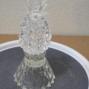 May include: A clear, faceted glass pineapple figurine featuring a textured, diamond-cut pattern on the fruit body and a pointed, crystalline leaf top. The object sits on a wide, flared, and scalloped glass base with a matching geometric design.
