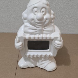 May include: A white ceramic clown figurine with a smiling face, a bow tie, and a hat. The clown is holding a rectangular frame with a dark interior. The figurine has a glossy finish.