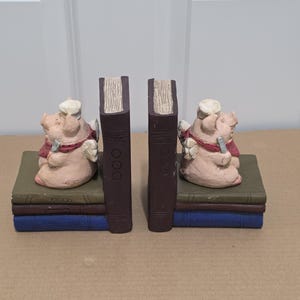 May include: A pair of bookends featuring pink pig figurines wearing white chef hats and red scarves. Each pig is positioned next to a vertical brown book spine, with the base of each bookend consisting of a stack of three books in green, brown, and blue.