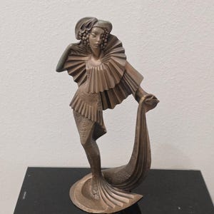 May include: A bronze-toned sculpture of a standing figure wearing a textured garment with a large, pleated ruff collar. The figure has a stylized headpiece and holds a long, flowing piece of fabric that extends down to the circular base.