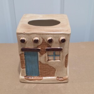 May include: A square ceramic tissue box cover designed to resemble a small adobe building. The exterior features a tan stucco texture with exposed brown brick details, a vertical blue door, a blue window, and four dark brown circular roof beams.