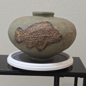 May include: A round, muted green ceramic vase with a wide, bulbous body and a narrow opening at the top. The front of the vase features a textured, dark brown relief carving of a fish, showing detailed scales and fins in a fossil-like style.