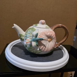 May include: A ceramic teapot with a hummingbird and flower design. The teapot has a white base with a pink flower, green leaves, and a brown handle. A colourful hummingbird is perched on the flower. The teapot sits on a black surface with a white rim.