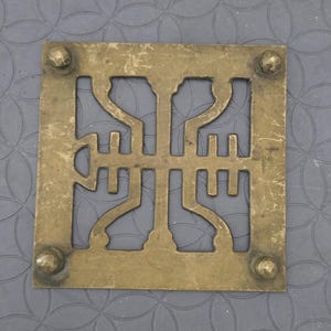 May include: Square brass trivet with an openwork design. The trivet has a geometric pattern in the center and rounded studs at each corner. The trivet is on a gray surface with a repeating circular pattern.