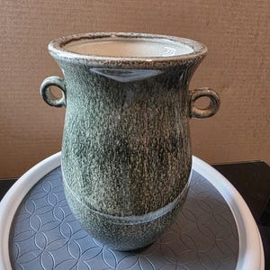 May include: A ceramic vase with a textured, mottled green and gray finish. The vase has a wide, slightly flared rim and two small loop handles. The interior is a lighter color, and the vase sits on a gray patterned surface.