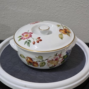 May include: A white porcelain lidded casserole dish featuring a delicate botanical pattern of pink flowers, green leaves, and orange fruit. The round vessel has two side handles and a gold-trimmed lid with a central knob handle.