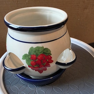 May include: A white ceramic strawberry pot with a blue rim and accents. The pot features a colorful strawberry design and two side compartments. It sits on a blue saucer and a gray patterned surface.