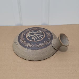 Puede incluir: Un recipiente de cerámica redondo y plano con un exterior de color beige claro texturizado y un panel superior circular de color marrón oscuro. La parte superior presenta un diseño de hojas grabado en el centro rodeado por un patrón de abanico. Un pico corto y ensanchado sobresale de un lado.