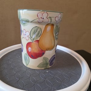 Pode incluir: Um recipiente cilíndrico de cerâmica com fundo creme, adornado com ilustrações pintadas à mão de frutas e flores. O design inclui uma maçã vermelha, uma pera amarela, ameixas roxas e flores brancas com folhas verdes. A borda é aparada com uma borda verde.