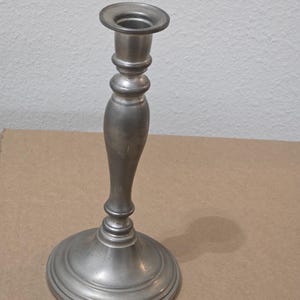 May include: A silver-colored candlestick with a round base and a slender, curved stem. The top has a circular holder for a candle. The metal has a slightly aged appearance, suggesting a vintage or antique style.