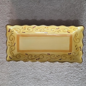May include: A rectangular serving platter with a scalloped edge. The platter is painted yellow with a light brown border and a central rectangular design. Decorative brown swirls and red berry accents adorn the edges.