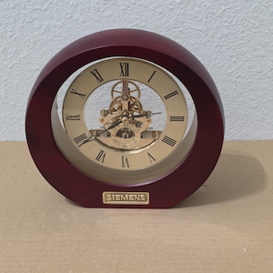May include: A round table clock with a dark mahogany-coloured wooden frame and a gold-toned face featuring black Roman numerals. The centre displays an exposed gold-coloured mechanical gear movement and a small seconds sub-dial. A gold plaque reads SIEMENS.