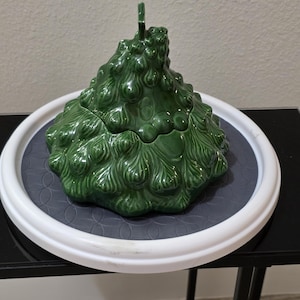 May include: A glossy, dark green ceramic lidded container shaped like a stylized Christmas tree. The surface features a textured, molded design resembling pine needles and branches, with a small star-shaped finial at the very top of the lid.
