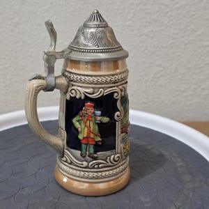 Könnte beinhalten: Ein dekorativer Bierkrug aus Keramik mit einem Klappdeckel aus Metall und einem Griff. Der Krug ist mit einer gemalten Szene einer Person in traditioneller Kleidung verziert. Die Farbpalette umfasst Beige, Blau, Grün und Rot.
