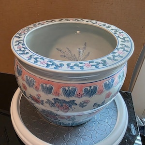 May include: A round ceramic planter featuring a white base with hand-painted floral and leaf motifs in shades of pink, blue, and green. The wide rim is decorated with a repeating vine pattern, and the interior base displays a painted fern design.