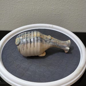 May include: A ceramic sculpture of a stylized animal with a rounded body and an elongated neck. The piece features a beige base color with vertical blue and grey drip-style glaze patterns, a small curled tail, and a smooth, glossy finish.