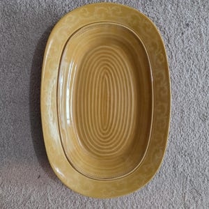 May include: Mustard yellow oval serving platter with a concentric swirl design in the center. The platter has a raised rim with a decorative pattern and a contrasting border. A stylish piece for serving food.