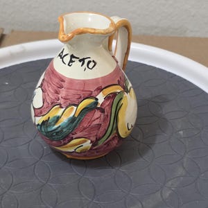 May include: A small, hand-painted ceramic pitcher with a handle and spout. The pitcher is white with a red and yellow floral design and the word "ACETO" in black script. The rim and handle are trimmed in orange.