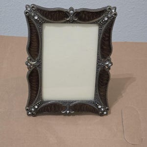 May include: An ornate rectangular photo frame with a dark brown wood-grain finish and silver-toned metal accents. The frame is embellished with small, clear rhinestones and decorative flourishes.