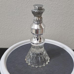 Könnte beinhalten: Ein dekorativer Flaschenverschluss mit einem klaren, facettierten Glassockel und einem polierten, reflektierenden silberfarbenen Metallaufsatz. Der Metallteil besteht aus einem gestuften, zylindrischen Design mit einem geriffelten Fächerdetail.