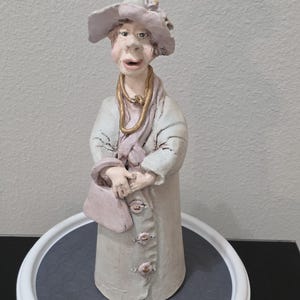 May include: A hand-sculpted clay figurine of a person wearing a wide-brimmed hat, a long pale coat with three buttons, and a scarf. The figure has a gold-colored necklace, holds a small handbag, and features a textured, matte finish in muted tones.