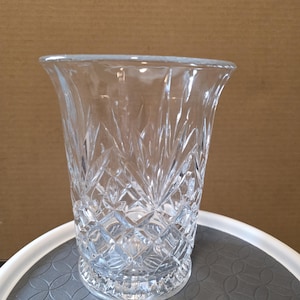 May include: A clear, cut-glass vase with a flared top and diamond and leaf pattern. The vase is approximately 7 inches tall and has a wide opening. The glass is transparent, allowing light to pass through, and is suitable for displaying flowers.