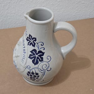 May include: A stoneware jug with a handle and spout. The off-white jug is decorated with blue grape and leaf designs, and the word "Rotfeller" is written on the side. The jug has a capacity of 0.5 litres.