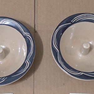 Può includere: Due pirottini per mele in ceramica. Ognuno ha un interno beige e un bordo blu scuro con un motivo a onde bianche. Un piccolo stelo cilindrico si alza dal centro di ogni pirottino.