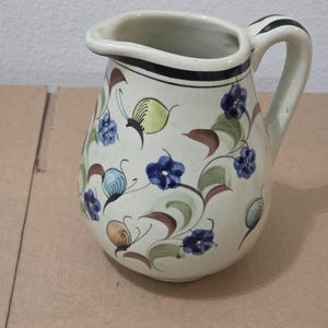 Peut inclure: Une cruche en céramique de couleur crème ornée d'un motif floral peint à la main avec des fleurs bleues, des feuilles vertes et des accents bruns. La cruche possède une anse incurvée à rayures noires et un liseré noir autour du col.