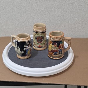 May include: Three ceramic beer steins with handles. The left stein is dark blue with a castle and figure. The center stein is light beige with a crest and the text Hannover. The right stein features a colorful scene of three figures sitting.