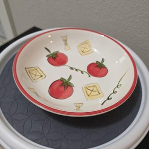 May include: A round, white ceramic bowl with a red rim. The bowl is decorated with hand-painted red tomatoes and yellow pasta shapes, connected by green vines. The bowl is sitting on a gray and white surface.