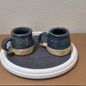 May include: Two handmade ceramic mugs featuring a rounded shape with a glossy dark blue glaze on the upper portion and an unglazed, matte beige finish on the lower base. Each mug includes a single curved handle attached to the side.