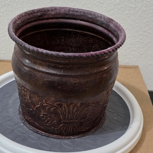 Pode incluir: Um vaso cilíndrico de metal castanho escuro com um acabamento desgastado e texturizado. O exterior apresenta um padrão floral em relevo à volta da base e um rebordo espesso semelhante a uma corda no topo. O interior é profundo e oco.