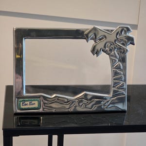 May include: A silver-colored rectangular picture frame with a palm tree design on the right side. The frame has a small rectangular label that reads "Can Cun" in green and white. The frame is sitting on a black table.