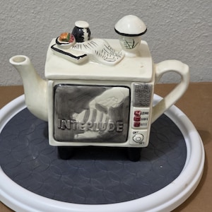 Könnte beinhalten: Eine cremefarbene Teekanne in Form eines Vintage-Fernsehers mit schwarzen Akzenten. Der Bildschirm zeigt das Wort "INTERLUDE". Auf der Oberseite befinden sich ein kleiner Teller mit Essen, ein kleiner schwarzer Behälter und ein pilzförmiger Deckel. Die Teekanne steht auf einem grau-weißen Tablett.