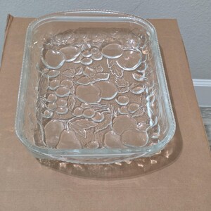 Puede incluir: Una fuente de horno rectangular de vidrio transparente con un patrón de relieve moldeado de varias frutas y hojas en la superficie inferior. El recipiente tiene esquinas redondeadas y un detalle de borde texturizado con cuentas en la parte superior.