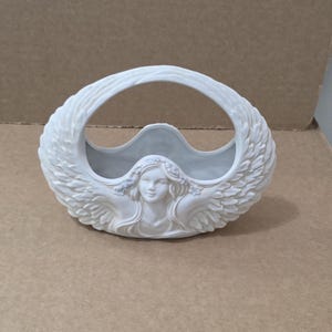 May include: A white ceramic basket featuring a sculpted figure of a person with long hair and outstretched wings that form the sides and handle of the vessel. The design includes detailed feather textures on the wings and floral accents in the hair.