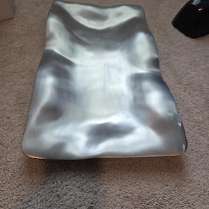 May include: A silver-colored, organically shaped decorative tray. The tray has a smooth, reflective surface with subtle curves and indentations. The tray's design is reminiscent of a natural form, adding a touch of modern elegance to any space.