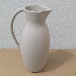 May include: A matte light grey ceramic pitcher with a smooth, rounded body, a curved handle on the left side, and a wide, open spout at the top. The vessel features a simple, uniform design with no additional patterns, textures, or decorative elements.