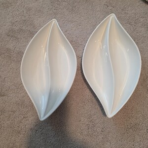 May include: Two white, leaf-shaped serving dishes. Each dish is divided into two sections, with a smooth, glossy finish. The dishes are placed side by side on a gray surface, showcasing their elegant design.