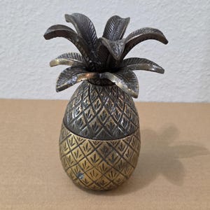 Puede incluir: Un recipiente metálico con forma de piña y tapa extraíble. El objeto presenta un patrón exterior texturizado de líneas entrecruzadas en tonos bronce antiguo y latón, rematado con una corona de hojas puntiagudas y detalladas en un tono oscuro.