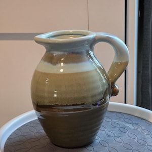 Puede incluir: Jarra de cerámica hecha a mano con cuerpo redondeado y asa curva. La jarra presenta un degradado de color, pasando de una base marrón oscuro a beige y azul claro en el borde. Un pequeño botón decorativo está en el lateral.