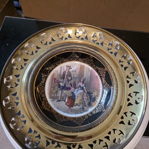 Könnte beinhalten: Ein dekorativer Teller mit einem goldfarbenen, durchbrochenen Rand und einem zentralen Bild einer historischen Szene. Der innere Teil des Tellers ist schwarz mit goldenen Details. Das Bild zeigt Figuren in historischer Kleidung. Der Teller steht auf einer weißen Oberfläche.