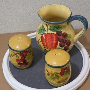 May include: A ceramic pitcher and two matching shakers featuring a yellow base with hand-painted red fruit, purple grapes, and green leaves. The pitcher has a curved handle and a blue rim, while the shakers have rounded tops with small holes.