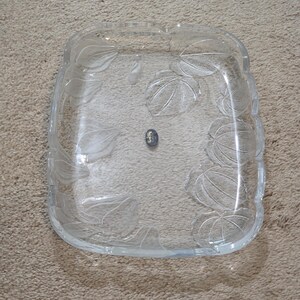 May include: A rectangular clear glass serving tray featuring a decorative embossed pattern of frosted leaves along the edges. The center of the tray is smooth and transparent, and a small dark oval sticker is affixed to the middle of the glass surface.