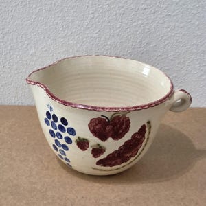 Può includere: Una ciotola in ceramica color crema con beccuccio e manico laterale. L'esterno presenta uva blu e fragole rosse dipinte a mano. Il bordo è decorato con una sottile linea testurizzata color marrone scuro, aggiungendo un dettaglio alla forma.