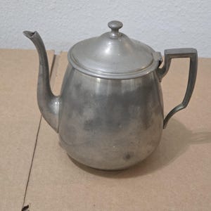 May include: A vintage silver-colored teapot with a lid and a handle. The teapot has a curved spout and a rounded body. The handle is rectangular. The lid has a small knob on top. The teapot is made of metal.