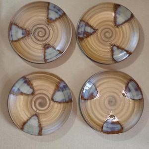 May include: A set of four round ceramic plates featuring a tan base with a spiral pattern. Each plate is decorated with three distinct triangular accents in shades of blue and brown, arranged symmetrically around the center of the plate surface.