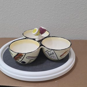 May include: A three-section ceramic serving dish with a white rim and a dark gray interior. The dish has a whimsical design with black outlines, yellow interiors, and a small figure on top. One section is labeled "Brie".