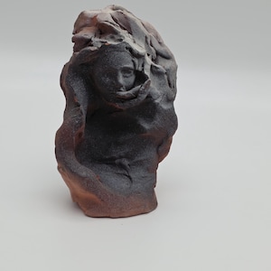 May include: A hand-sculpted ceramic bust featuring a dark, textured, charcoal-colored face emerging from a raw, reddish-brown clay form. The sculpture has an uneven, organic surface with deep shadows and a matte finish, capturing a human-like visage.